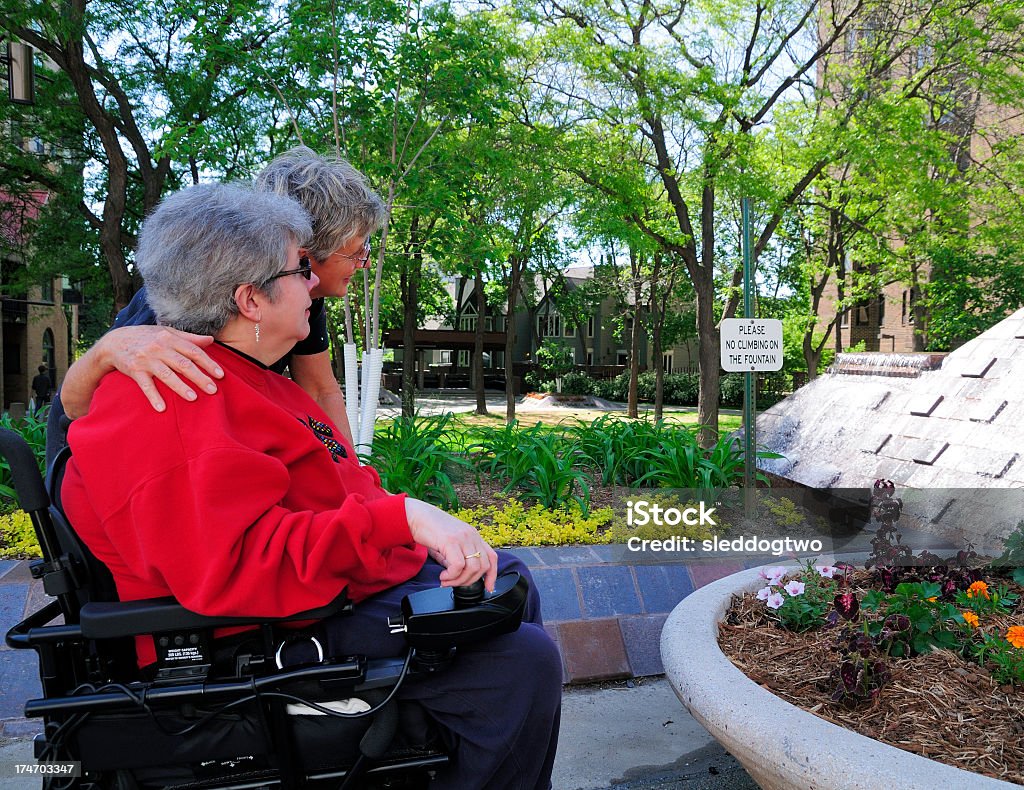 A woman in a wheelchair accompanied by another woman Two middle aged women in an urban park on a nice day, one has arm around the other who is seated in a power wheelchair. Multiple Sclerosis Stock Photo