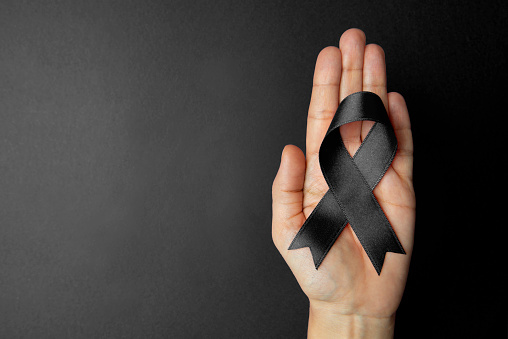 Directly above view of a hand holding black awareness ribbon over black background. Some issues for what the black awareness ribbon stands for, anti-terrorism, gun control, mourning, narcolepsy, melanoma, sadness, remembrance,POW / MIA, 9/11 Loss Remembrance, trauma and sleep disorder.