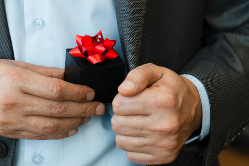 Chest view of an unrecognizable businessman wearing a suit who is about to give a gift box out of his jacket. The black gift box is wrapped with red ribbon.