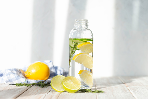 Bottle of infused water on white wood with a slice of lemon , cucumber and rosemary leaf in it.