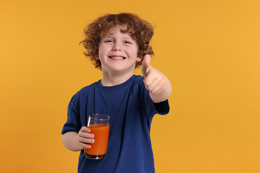 Cute little boy with glass of fresh juice showing thumb up on orange background