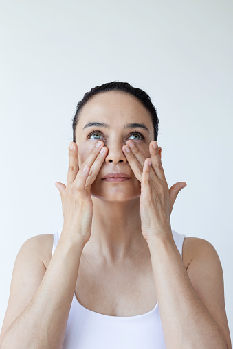 Caucasian female is doing face yoga gymnastics for non-surgical rejuvenation and is looking at camera in front of white background. Anti aging concept.