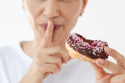 Caucasian woman is eating doughnut with sprinkles in front of white background. She is doing silence gesture with her finger in front of her mouth.