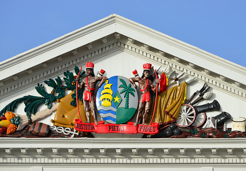 Suriname, Paramaribo: Suriname's emblem in the tympanum of the Presidential Palace - an 18th century building - the coat of arms of Suriname consists of an oval divided shield with a sailing ship at sea on the left half, and a royal palm on the earth on the right half. In the center there is a green diamond with a five-pointed star in it. The shield is flanked by two brown natives with bows and arrows, with a pennant underneath with the Latin motto 'Justitia, Pietas, Fides' (Justice, Piety, Fidelity). Pediment framed by scroll modillions.