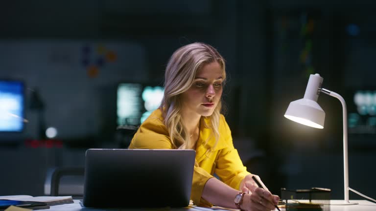 Night, office and woman with research for report, article and writing notes for news, blog and review. Reading, laptop and analysis of online information, update and writer working late in workplace