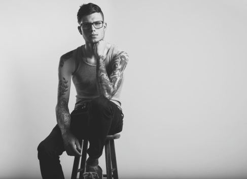 Studio portrait of a young caucasian tattooed urban hipster man. He is wearing glasses and a gray wife beater with jeans and a leather belt. Shot on a blank white background in a studio. Captured September 2012.