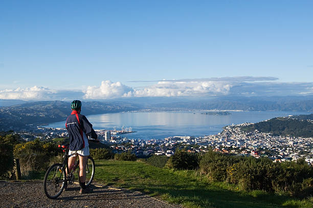 Mountain biker at summit "Mountain biker pauses at the summit of a hill, looking down to the harbor city below. Clear blue skies with a bank of puffy clouds on the horizon." mountain biking photos stock pictures, royalty-free photos & images