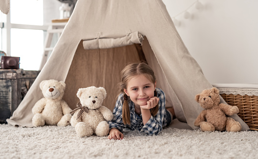 Little girl lying in wigwam with plush teddies in light kids room