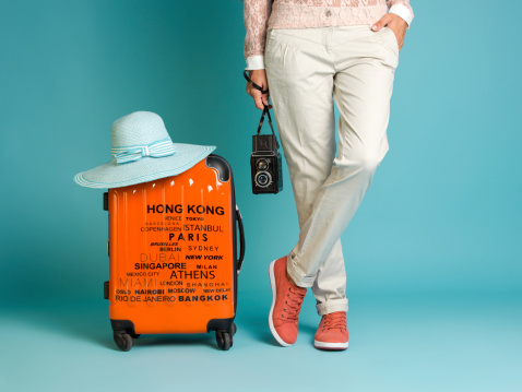 A young woman standing with a an orange suitcase beside her. She is wearing a cream colored pants and red tennis shoes with white soles. She is holding a twin lens reflex camera in right hand. Her left leg is crossed over her left leg. The woman's body is only visible from the waist down. Image taken in studio over blue background. 