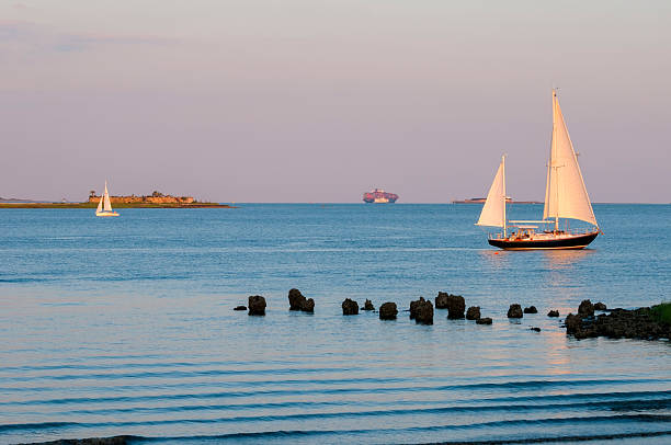 Two sailboats going opposite directions on an early summer evening in Charleston Harbor in South Carolina. In the far distance is a cargo ship and the Fort Sumter ferry. Fort Sumter is behind the sailboat on the right.