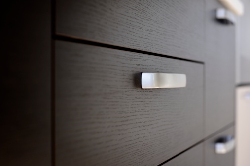 Close-up shot of dark-colored kitchen drawers with silver handles.  A white countertop is visible above the drawers, and there are light-colored appliances in the background.