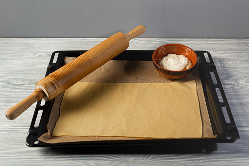 Rolling pin and baking sheet with dough and filling sits in a black oven tray on gray wooden table