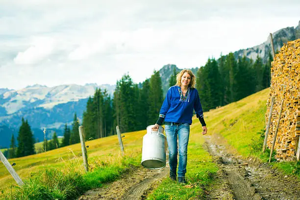 a young woman is carrying a milk canister. the simmental valley can be seen in the background.
