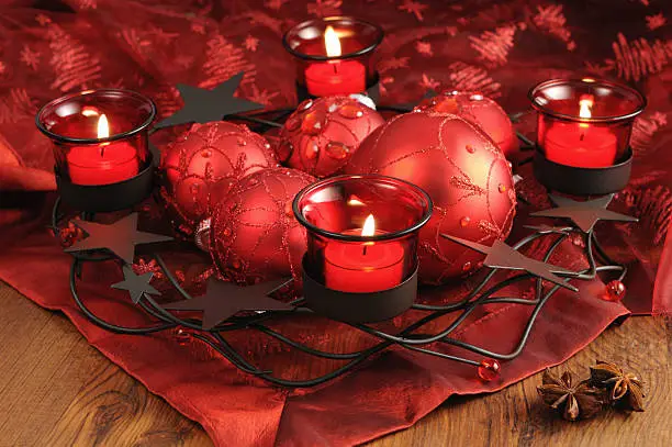 On table standing metal advent wreath with three red burning tealights and Christmas ornaments. In front two star anise on red tablecloth. Christmas background.