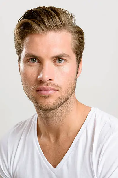 "Handsome young man headshot portrait. Stubbly beard on a strong jawline, a cool slicked back quiff and eye contact looking to camera. Neutral expression. White v-neck t-shirt.Handsome young man headshot portrait. Stubbly beard on a strong jawline, a cool slicked back quiff and eye contact looking to camera. Neutral expression. White v-neck t-shirt."