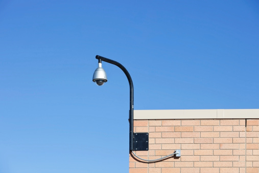 A 360 degree coverage surveillance camera is mounted on a pole to the side of a brick building against a clear blue sky.