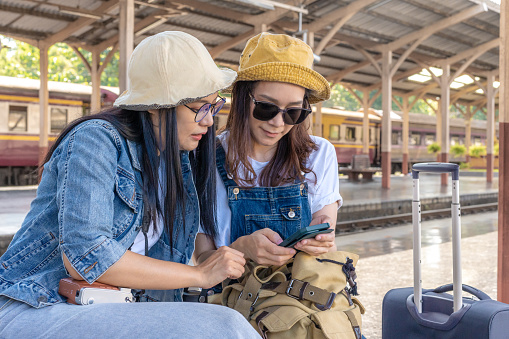 Two Asian female traveller friends sitting at a train station searching for their destination's direction on their phone