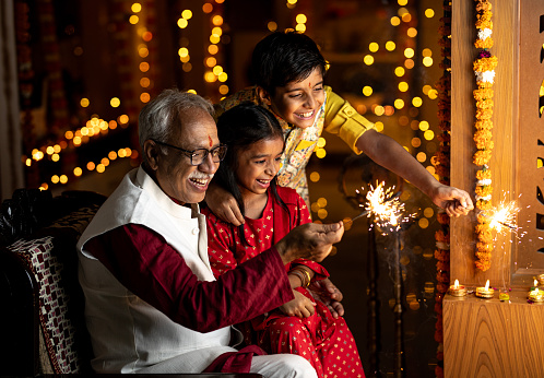 Cheerful grandfather and grandchildren playing with sparklers while celebrating Diwali festival at illuminated home