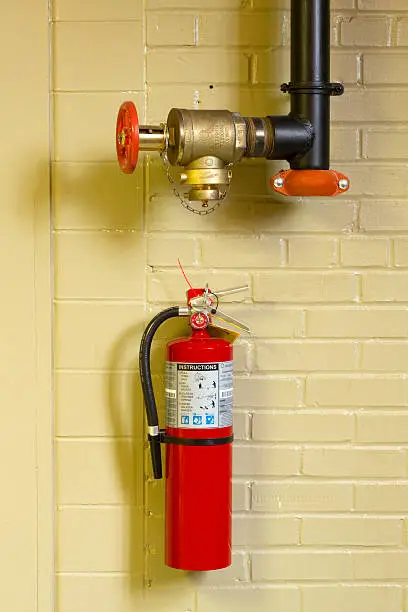 Shown here is a fire standpipe hose valve and a red fire extinguisher on a painted brick interior wall in a public building.