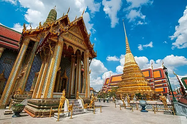 "Prasat Phra Thep Bidon in Grand Palace and Wat Phra Kaew Temple interior, Bangkok, Thailand. The Emerald Buddha temple. Visible are two of the many Buddha temple and the Golden Pagoda in interior of Grand Palace. Dramatic cloudscape with blue sky and cumulus clouds over the Grand Palace.See more images like this in:"