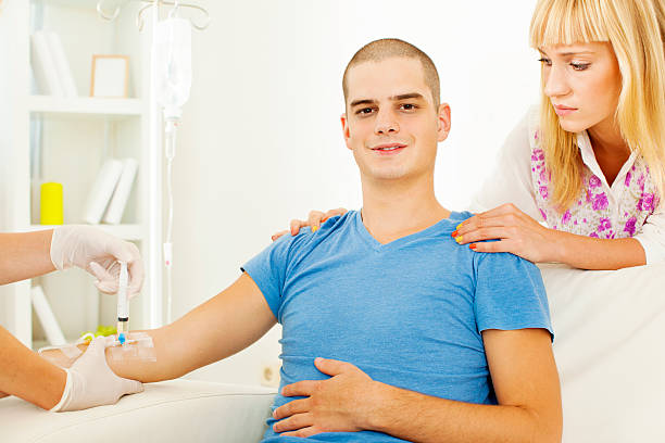 Young Man having Chemotherapy at home Portrait of an young man having chemotherapy therapy at home. Professional healthcare nurse preparing therapy while his woman standing behind him and touching his shoulders. Patient looking at camera with smile. Prime IV Hydration TherapyWhy are young people loving it? stock pictures, royalty-free photos & images