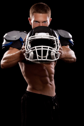 Young muscular football player posing with shoulder pads and holding protective helmet.