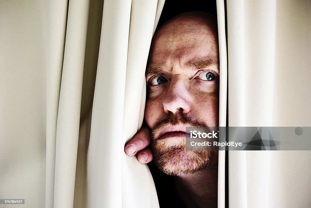 Suspicious bearded man peers through curtains fearfully "Peeping to the side through closed drapes, this man frowns worriedly; perhaps there is a real threat, perhaps it is all in his mind." Frowning Stock Photo