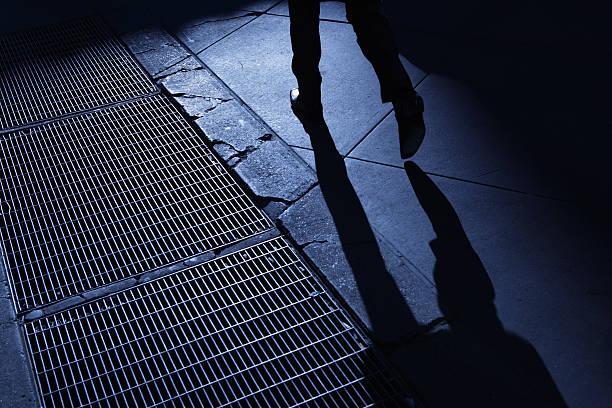 Shadows elongate across a city sidewalk as the sun sets, casting long lines against the pavement. Two feet in sneakers walk along the path, creating a dynamic interplay between light and dark. A metallic tactile paving line and bicycle wheel add urban detail.