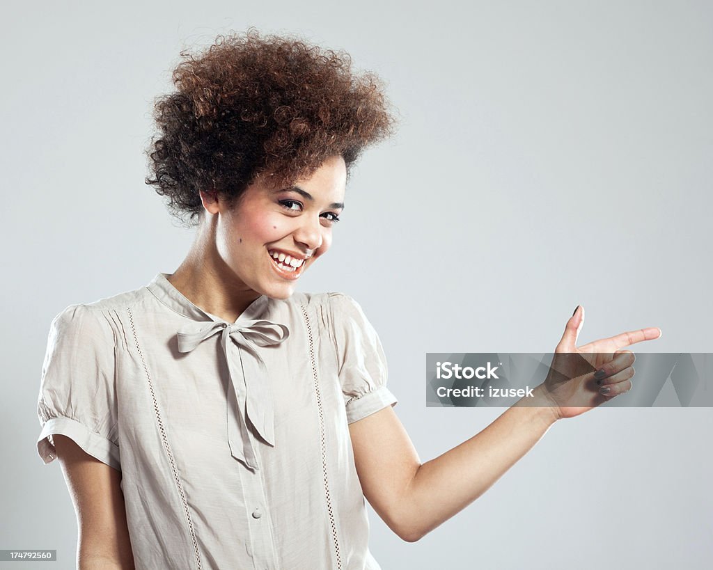Cute girl pointing at copy space Portrait of beautiful teenaged afro girl pointing with index finger at the copy space and smiling at the camera. Studio shot, grey background. Gray Color Stock Photo