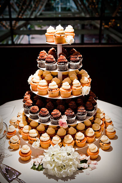 A tower of cupcakes at a wedding in place of the traditional wedding cake.  There are a mixture of vanilla chocolate chip with vanilla frosting and chocolate with chocolate frosting.  There are white and pink roses decorating the cupcake stand.  There is a beautiful white floral arrangement on the round table.