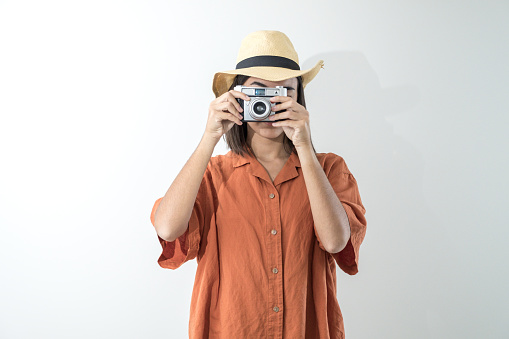 Happy young girl in hat takes photo with old camera in studio. Tourist in ochre or orange shirt on sightseeing trip.