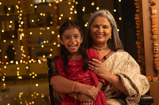 Portrait of cheerful senior woman and girl celebrating Diwali festival together at illuminated home