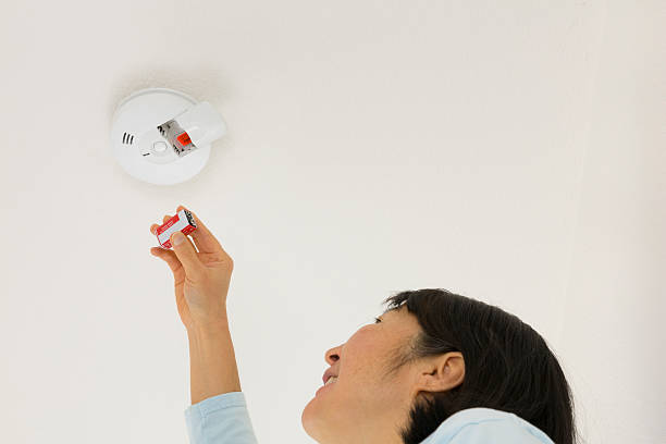Woman inserting smoke alarm battery A middle-aged Asian woman inserting a 9V battery into a ceiling-mounted smoke detector. smoke detector photos stock pictures, royalty-free photos & images