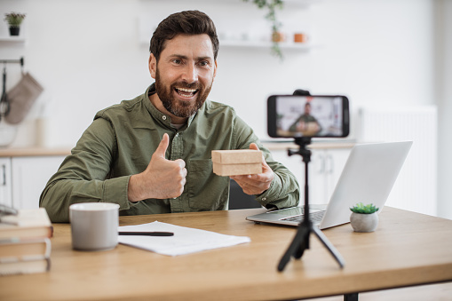 Emotional young guy opening cardboard box and looking inside with excitement while filming video on smartphone showing thumb up