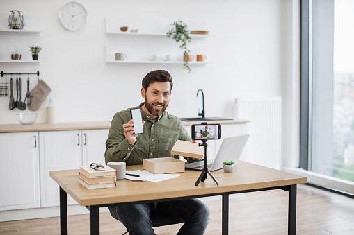 Positive handsome man recording video blog while unboxing new modern smartphone indoors. Interesting influencer leaving review about product and sitting at table with laptop in room.