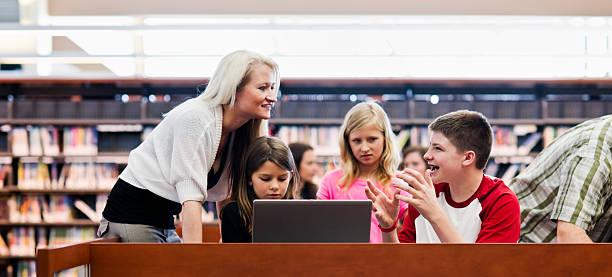 Group of children around laptop talking to adult in library stock photo