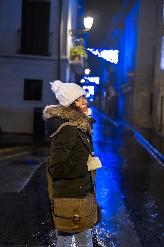 Young girl in coat, hat and gloves poses on an empty street in a village with blue lights. Navarra.