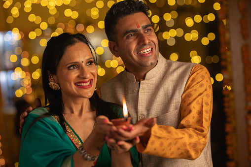 Smiling young husband and wife dressed in traditional clothing holding diya while celebrating Diwali festival at home