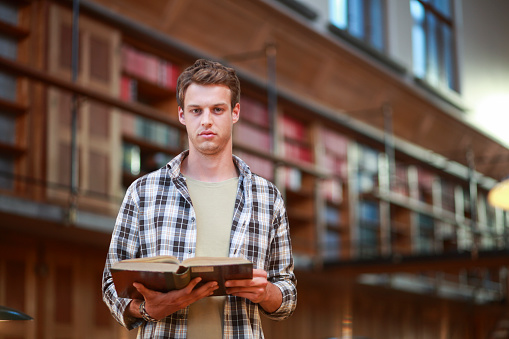 Young student holding a book and standing in the middle of a public library.