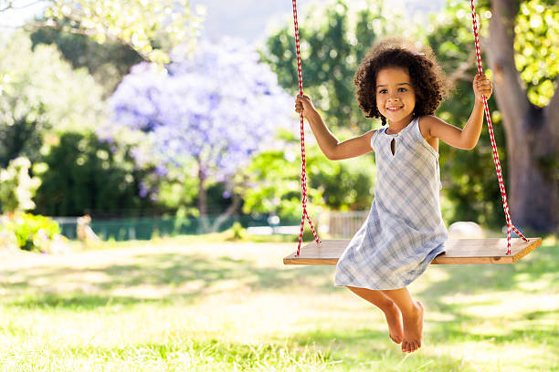 Smiling young girl on a swing in a park A young dark-skinned little girl wearing a white and blue dress and sitting on a wooden bench swing, smiling for the camera. There is grass below her. There are trees and a clear blue, sunny sky behind her. swing girl dress stock pictures, royalty-free photos & images