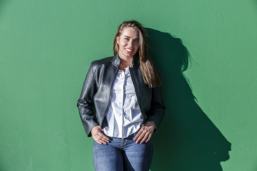 Confident young smiling female wearing leather jacket and jeans putting fingers in pockets while standing against green wall with shadow from sunlight