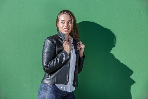 Positive young female adjusting black leather jacket while standing against green wall with shadow from sunshine on street and looking away