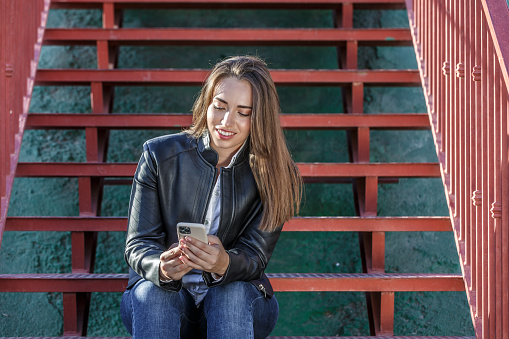 Young attractive female with long fair hair wearing leather jacket sitting on red staircase and chatting on social media via modern mobile phone during free time