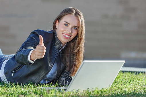 Satisfied young female remote worker in leather jacket lying on grass using laptop while showing thumb up gesture during online work outdoors
