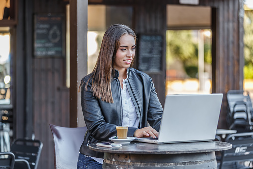 Positive young female freelancer in black leather jacket sitting at table with coffee cup and browsing netbook while working on remote project in outdoor cafe