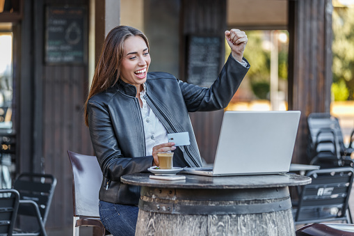 Excited young woman in leather jacket sitting at table in street cafe looking at laptop screen clenching and raising fist up while celebrating successful online bet holding plastic card