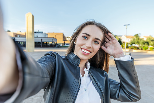 Pretty young woman with blonde hair and blue eyes, smiling while taking a selfie with her camera on the street.