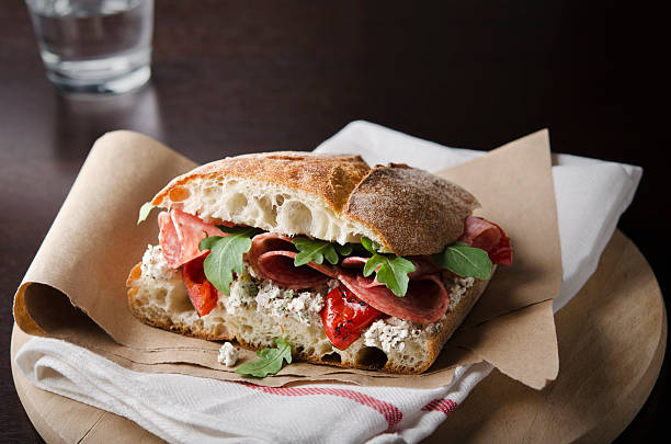 Artisan salami sandwich over a napkin in a round wood tray Rustic sandwich of salami, roasted red peppers, arugula, and herbed goat cheese on ciabatta bread. Shallow DOF. Ciabatta Roll stock pictures, royalty-free photos & images