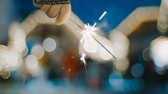 Hand of woman in gloves holding burning sparkler outdoors during festive night
