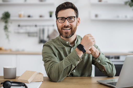 Mobile web camera view of cheerful guy in eyeglasses trying on new smart watch and recording video blog. Technology blogger making online review of modern gadget for social media followers.